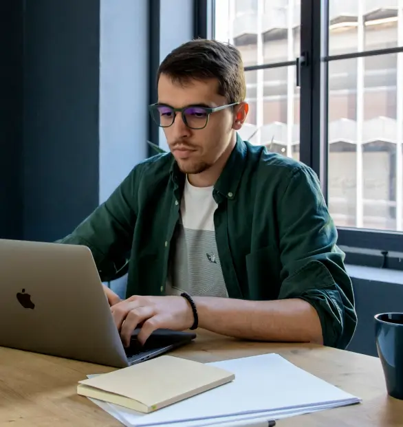 A focused man wearing glasses and a green shirt working on a MacBook at a wooden desk with a notebook, papers, and a cup nearby in a modern office setting.