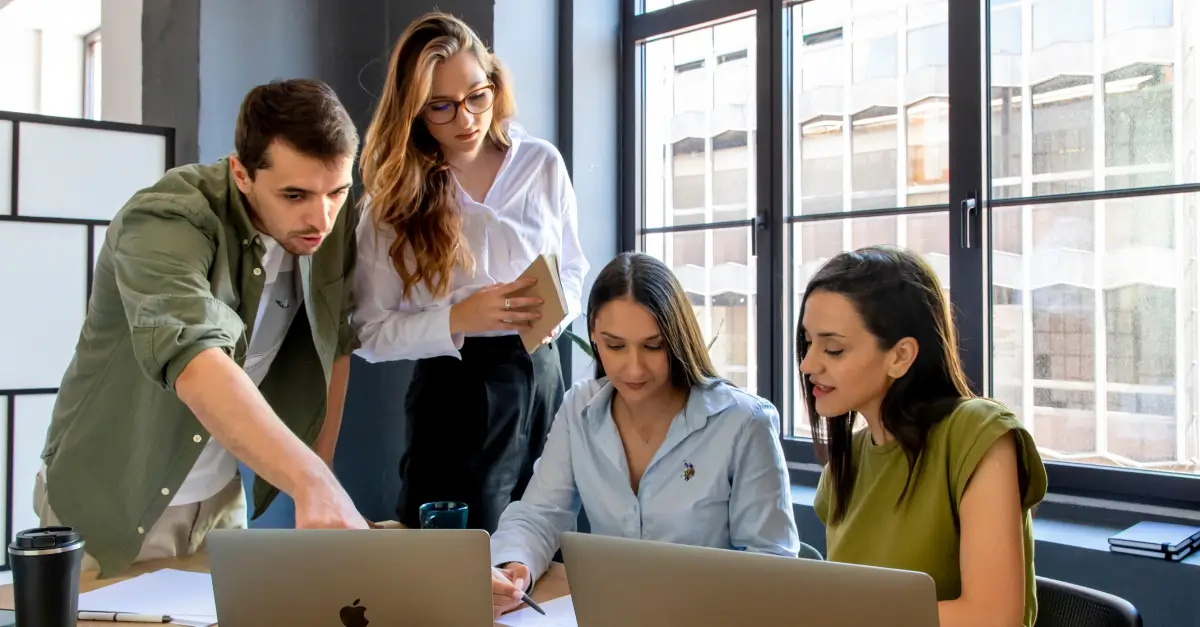 A group of four colleagues collaborating at a desk in a modern office, with one person pointing at a laptop screen while others engage with their devices and notes. They are working together, with large windows in the background letting in natural light.