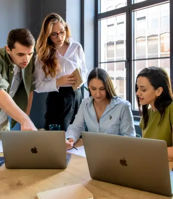 A group of four colleagues collaborating at a desk in a modern office, with one person pointing at a laptop screen while others engage with their devices and notes. They are working together, with large windows in the background letting in natural light.