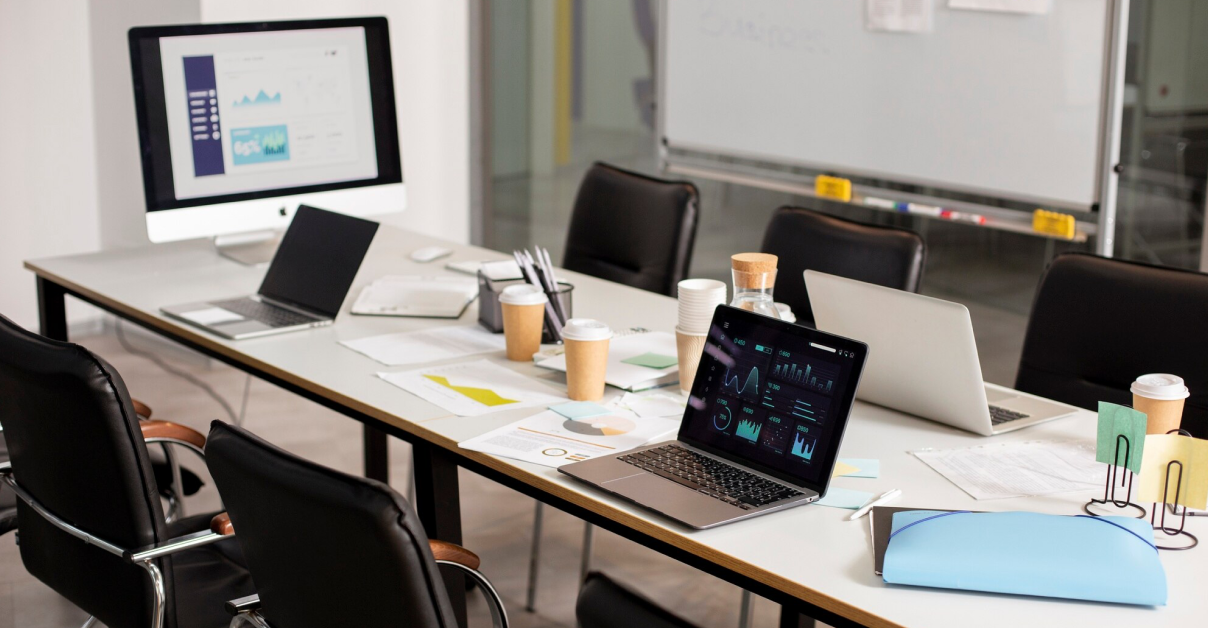 A conference room with a table set up for a meeting, featuring laptops displaying charts and graphs, documents, coffee cups, and a monitor showing data presentations.