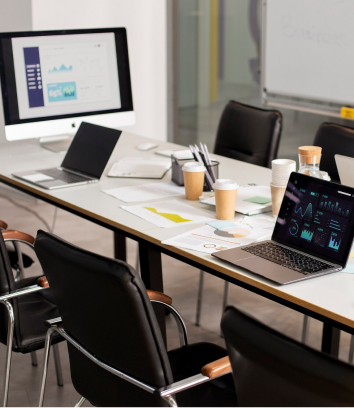 A conference room with a table set up for a meeting, featuring laptops displaying charts and graphs, documents, coffee cups, and a monitor showing data presentations.